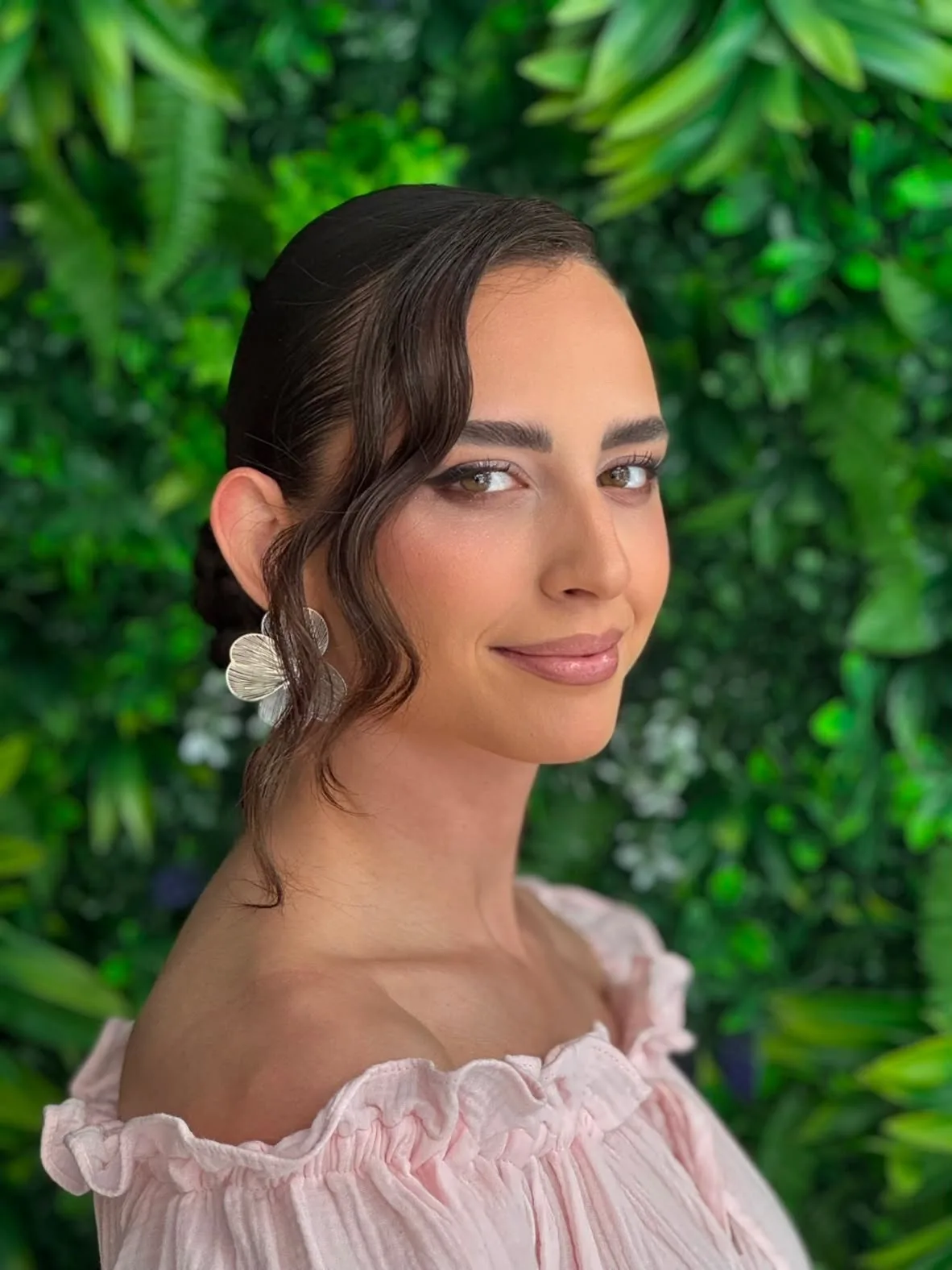 Romantic bridal makeup with warm bronze shadow, softly defined lashes and a glossy rose lip, photographed against a green foliage wall.