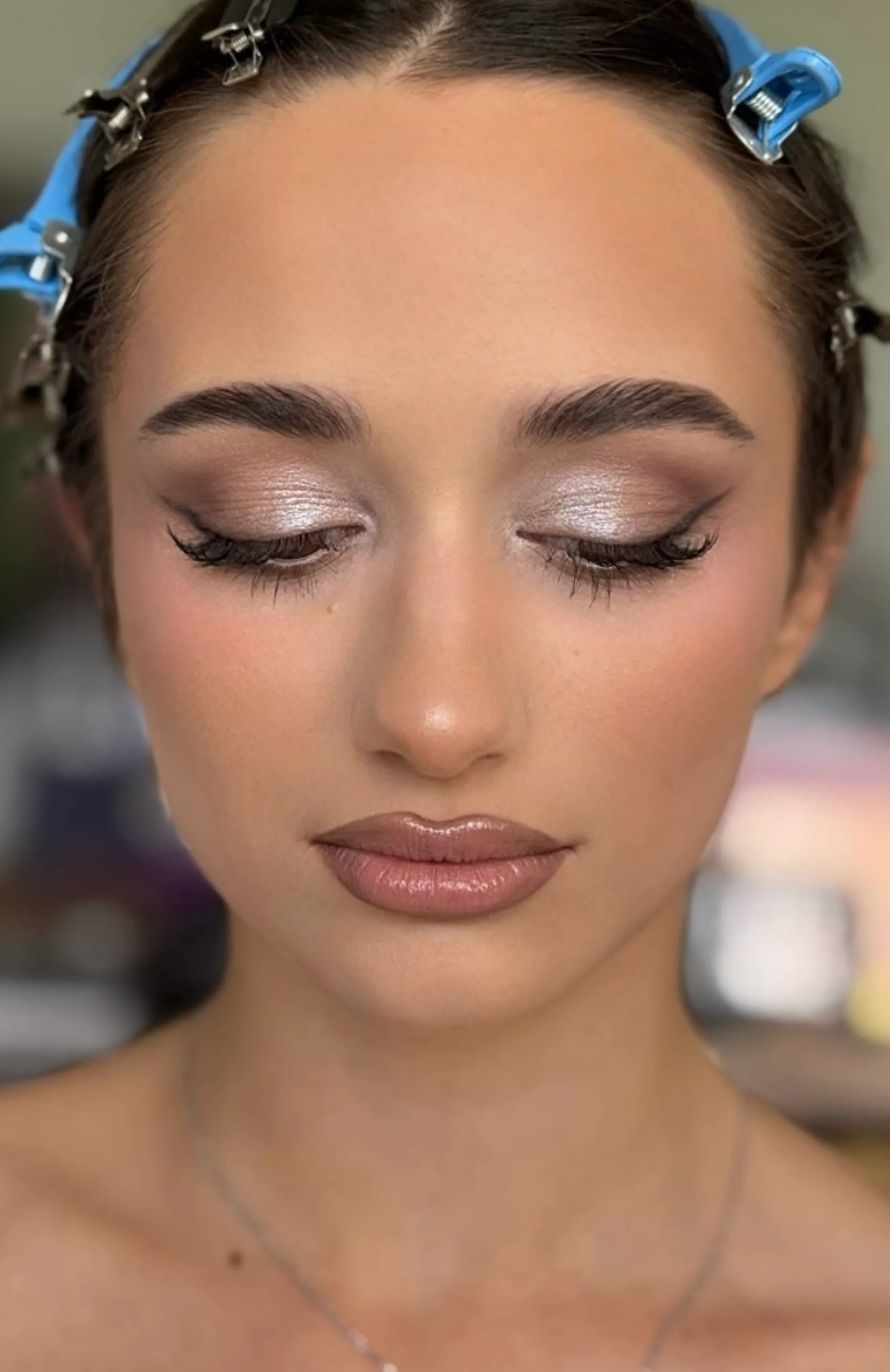 Eyes-closed detail of a satin champagne lid, feathered brows and a warm mauve lip, sectioning clips still in the hair.